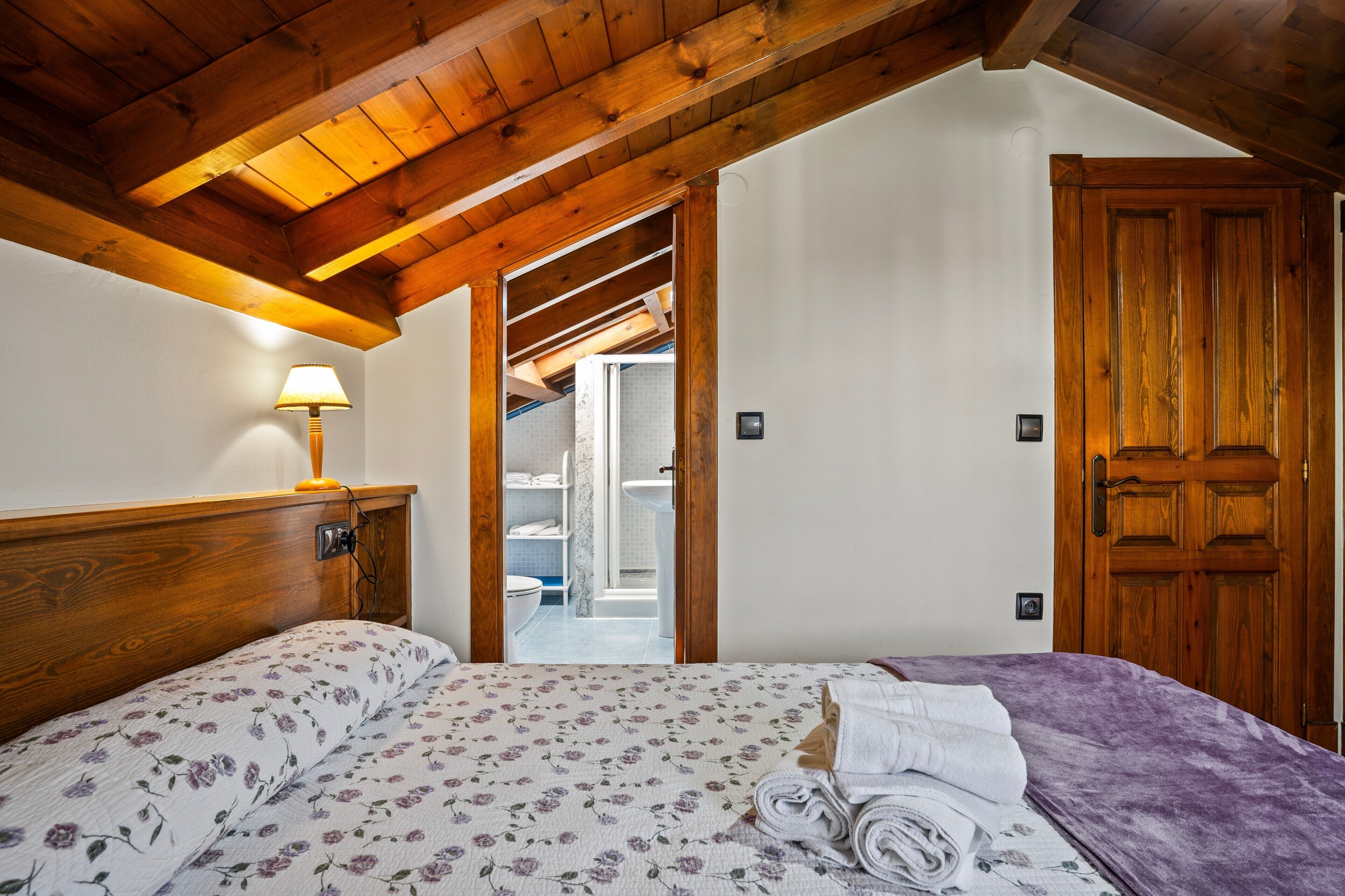 Een slaapkamer met een houten plafond, een bed met bloemenbedrukking, een nachtkastje met een lamp en een houten deur.