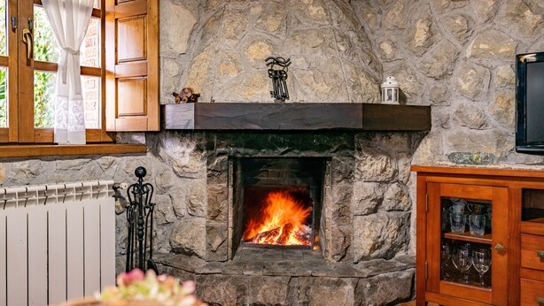 Una chimenea de piedra con fuego, contraventanas de madera, un radiador y un aparador con cristalería.
