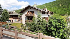 Un chalet à deux étages doté d’un balcon, entouré de verdure et de montagnes.