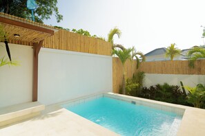 Una piscina con muelle de madera y pérgola, rodeada de una valla de bambú y plantas tropicales.