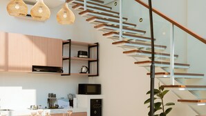 Una sala de estar moderna con un sofá, una mesa de comedor dispuesta con platos y vasos, una escalera con barandilla de cristal y lámparas colgantes.