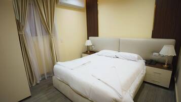 A neatly made bed with white linens, a bedside table with a lamp, and a wall-mounted air conditioner.