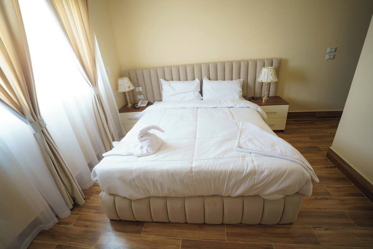 A neatly made bed with a headboard, two bedside tables with lamps, and a window with curtains.
