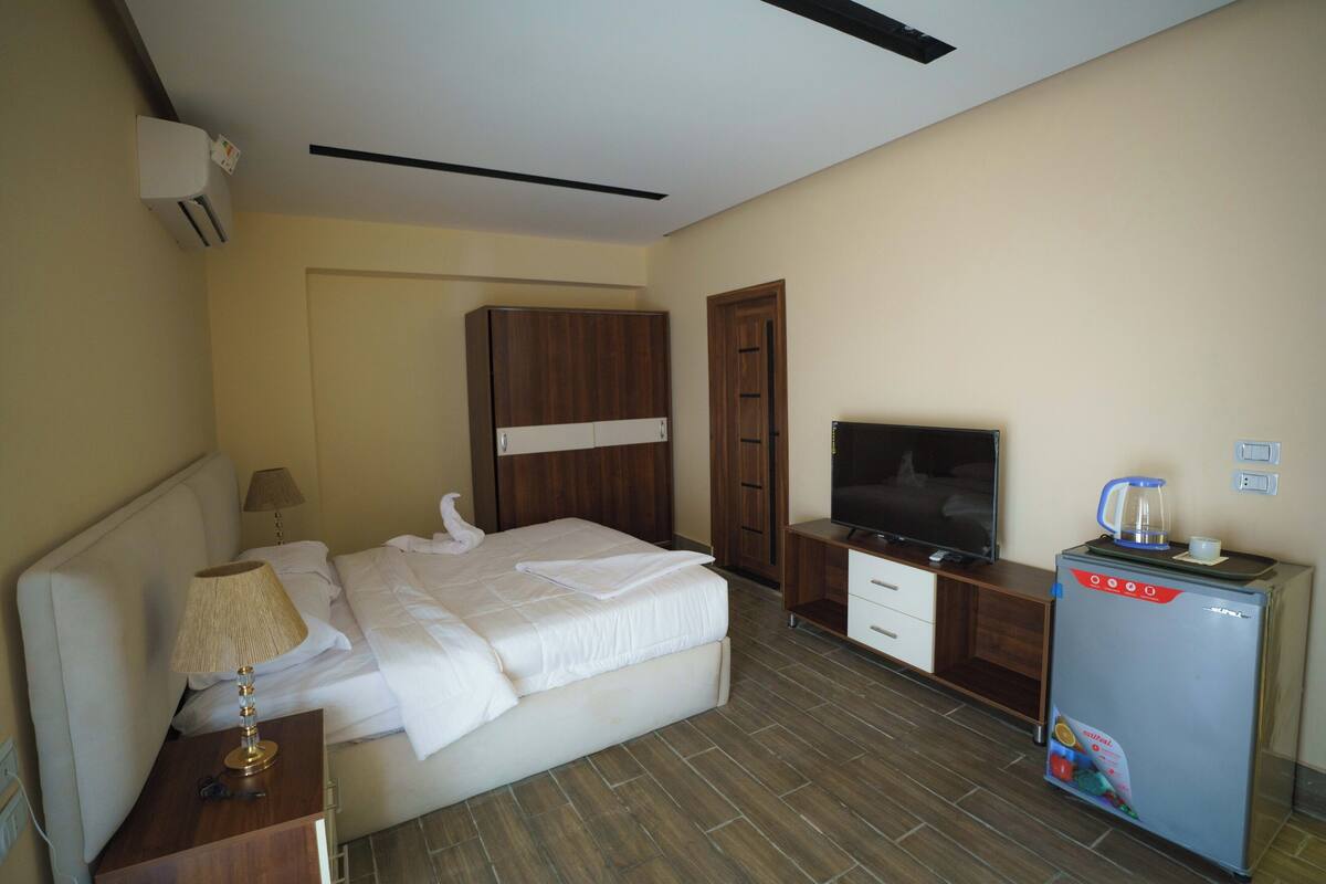 A hotel room with a bed, bedside table, lamp, wardrobe, TV, and a refrigerator.