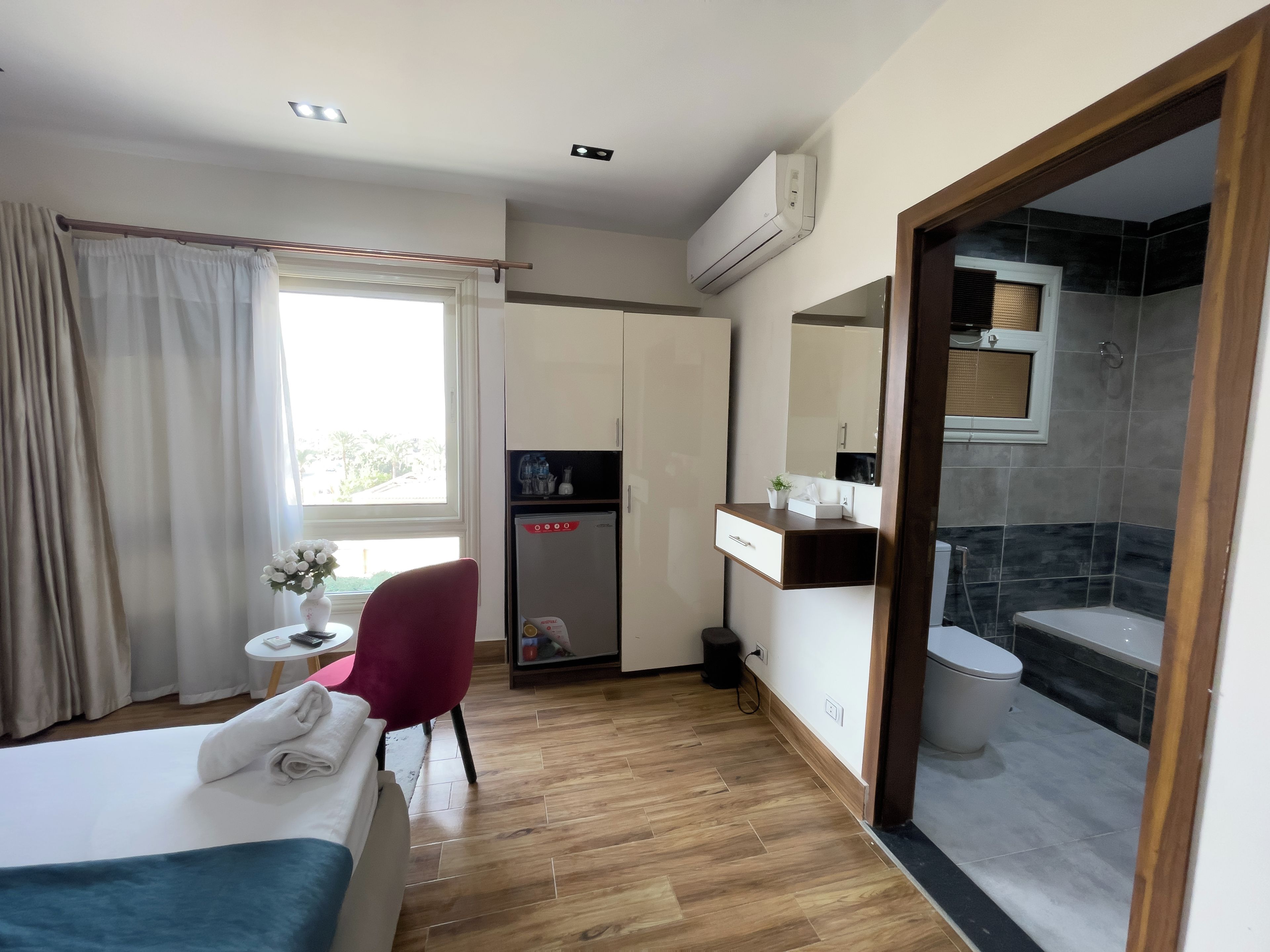 Habitación de hotel con cama, escritorio, silla, pequeña mesa con un florero, espejo, armario, baño con ducha y equipo de aire acondicionado.
