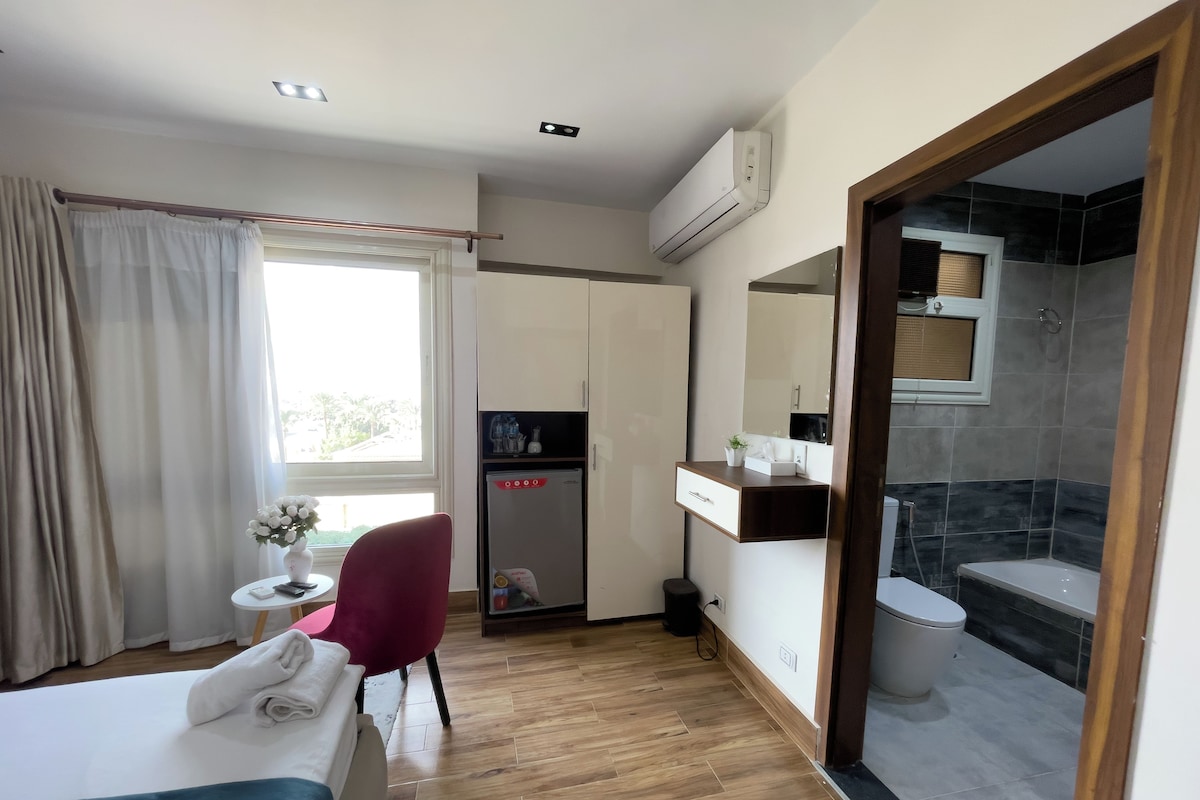 Ein Hotelzimmer mit Bett, Schreibtisch, Stuhl, einem kleinen Tisch mit Vase, Spiegel, Kleiderschrank und einem Badezimmer mit Dusche sowie einer Klimaanlage.
