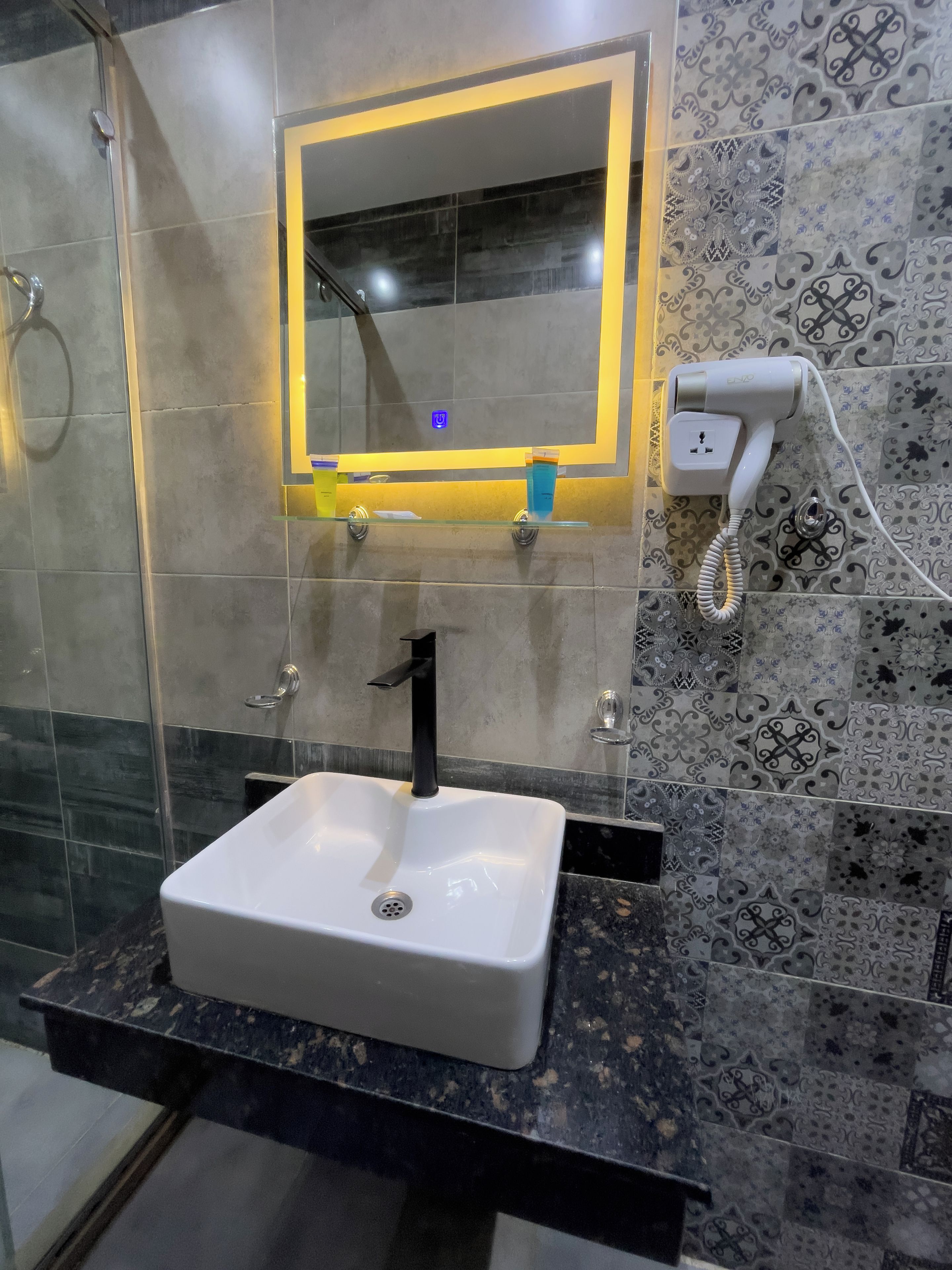 Un baño con un lavamanos blanco, un grifo negro, un espejo con luz amarilla y un secador de pelo enchufado.