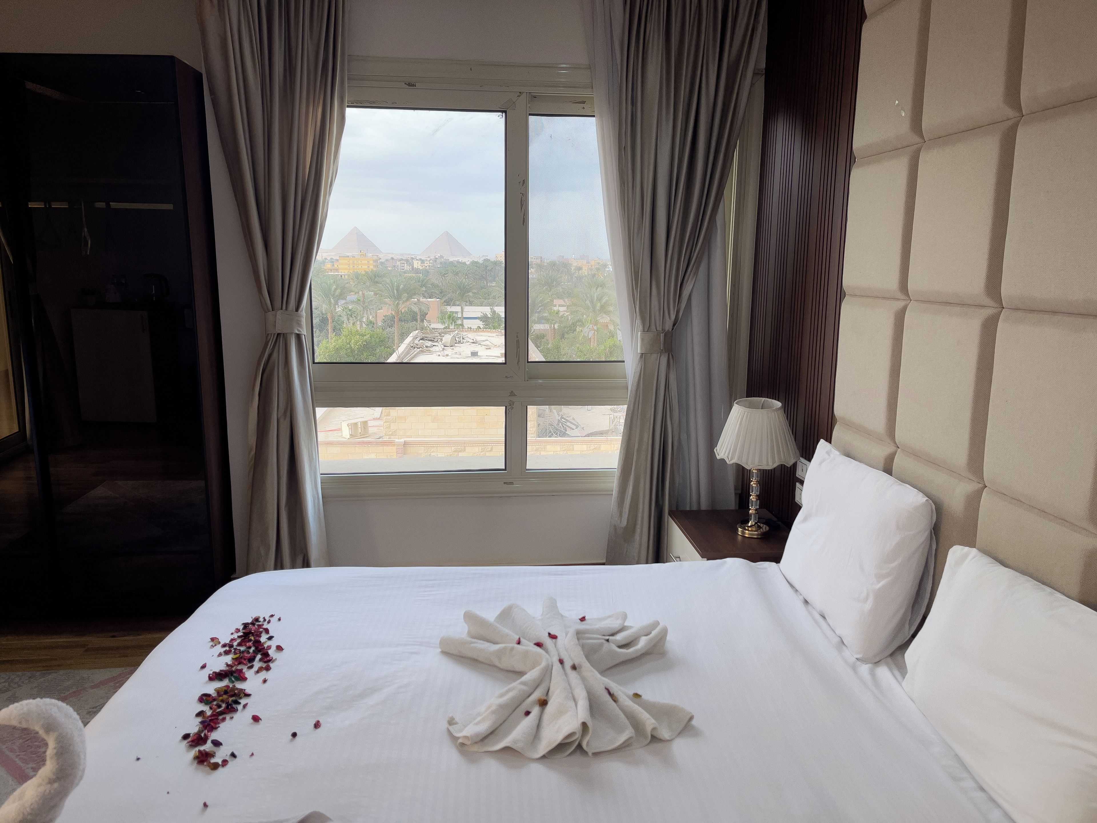 A hotel room with a bed, a nightstand with a lamp, a view of pyramids, and curtains.