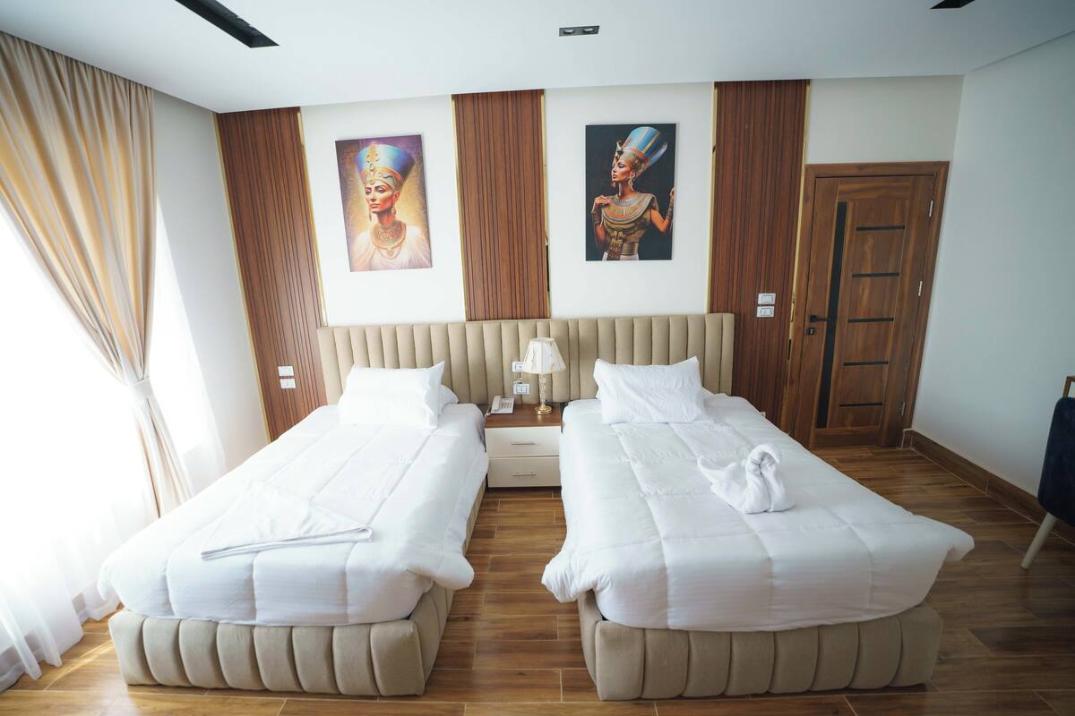 A hotel room with two beds, wooden flooring, a large window with curtains, and two wall-mounted paintings.