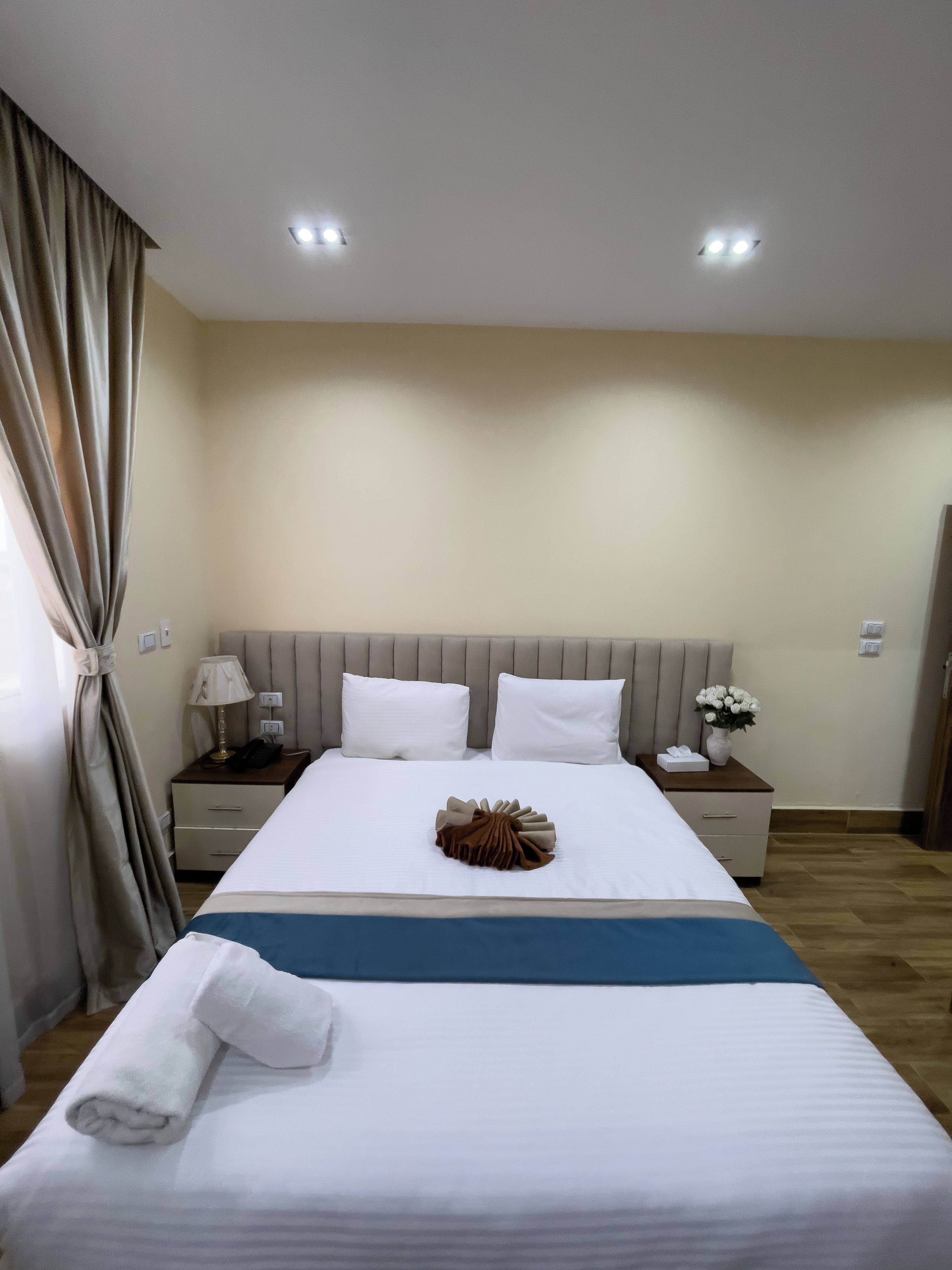 A hotel room with a bed, a nightstand, a lamp, a vase with flowers, and a curtain.