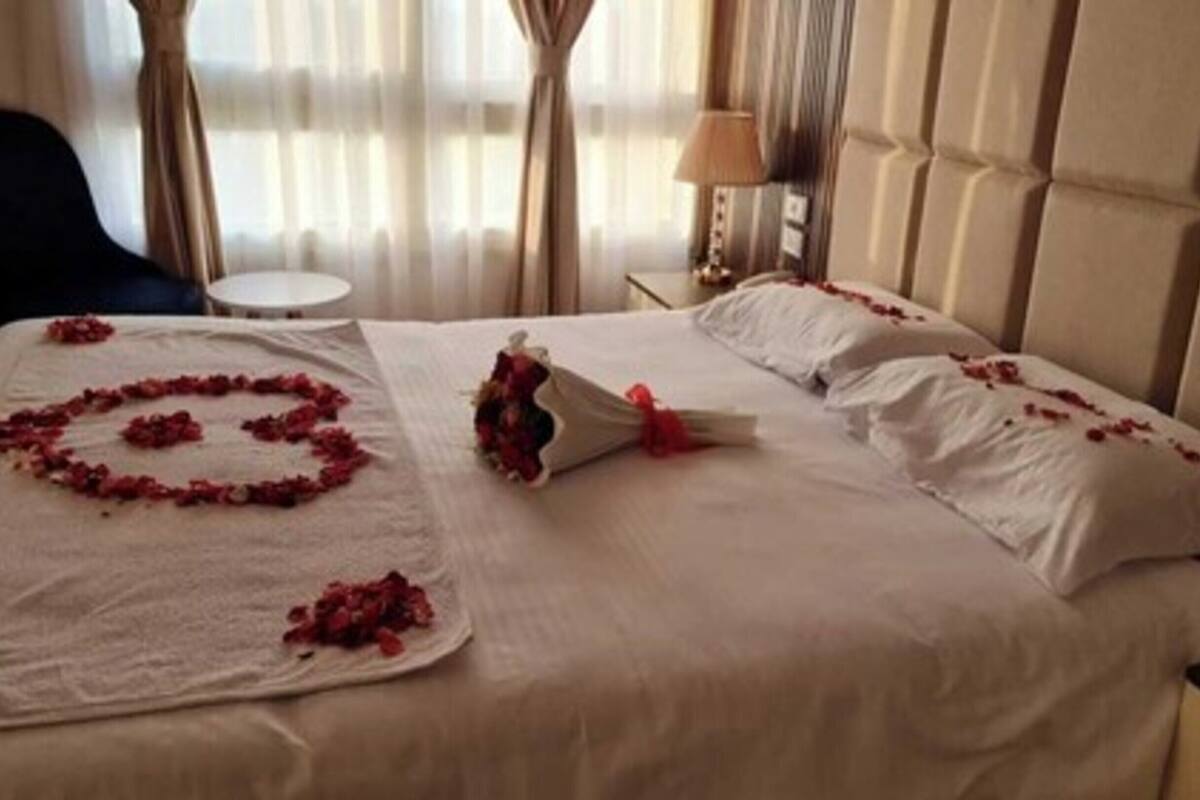 A hotel room with a bed adorned with rose petals arranged in a heart shape, a bouquet of flowers, and a small table with a lamp.
