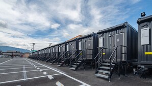 A row of black modular units with numbered doors, arranged in a parking lot with marked spaces and steps leading to each unit. - HOTEL R9 The Yard Yamanashichuo (Chuo)