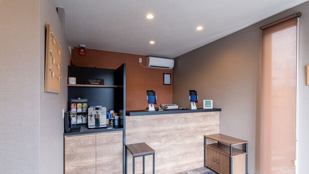A modern reception area with a wooden counter, two coffee machines, a small table, and a wall-mounted air conditioner. - HOTEL R9 The Yard Yamanashichuo (Chuo)