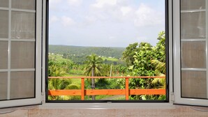 A window with a view of a lush green landscape, including palm trees and distant buildings. - Charmant Chalet Créole Avec sa Piscine Privée (Goyave)