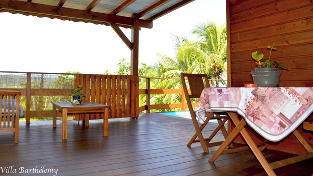 A wooden deck with a table and chairs, a covered area, and a view of palm trees. - Charmant Chalet Créole Avec sa Piscine Privée (Goyave)