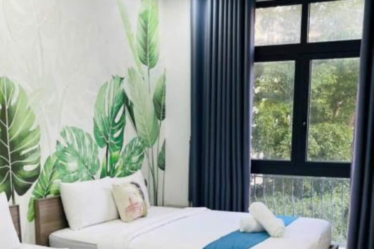 A hotel room with two beds, a large window with curtains, and a tropical leaf mural on the wall.