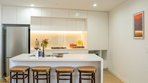 A modern kitchen with white cabinetry, a stainless steel refrigerator, a central island with stools, and a small countertop with a vase of flowers. - Executive Rental - Campbelltown (Campbelltown)