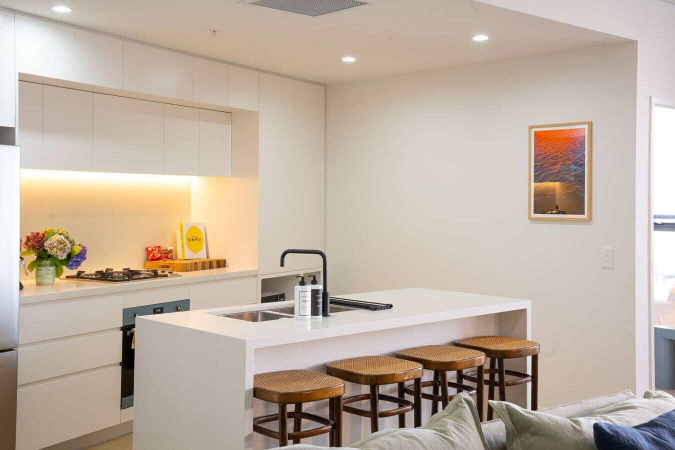 Uma cozinha moderna com armários brancos, uma ilha central com banquetas, uma pia e um fogão.