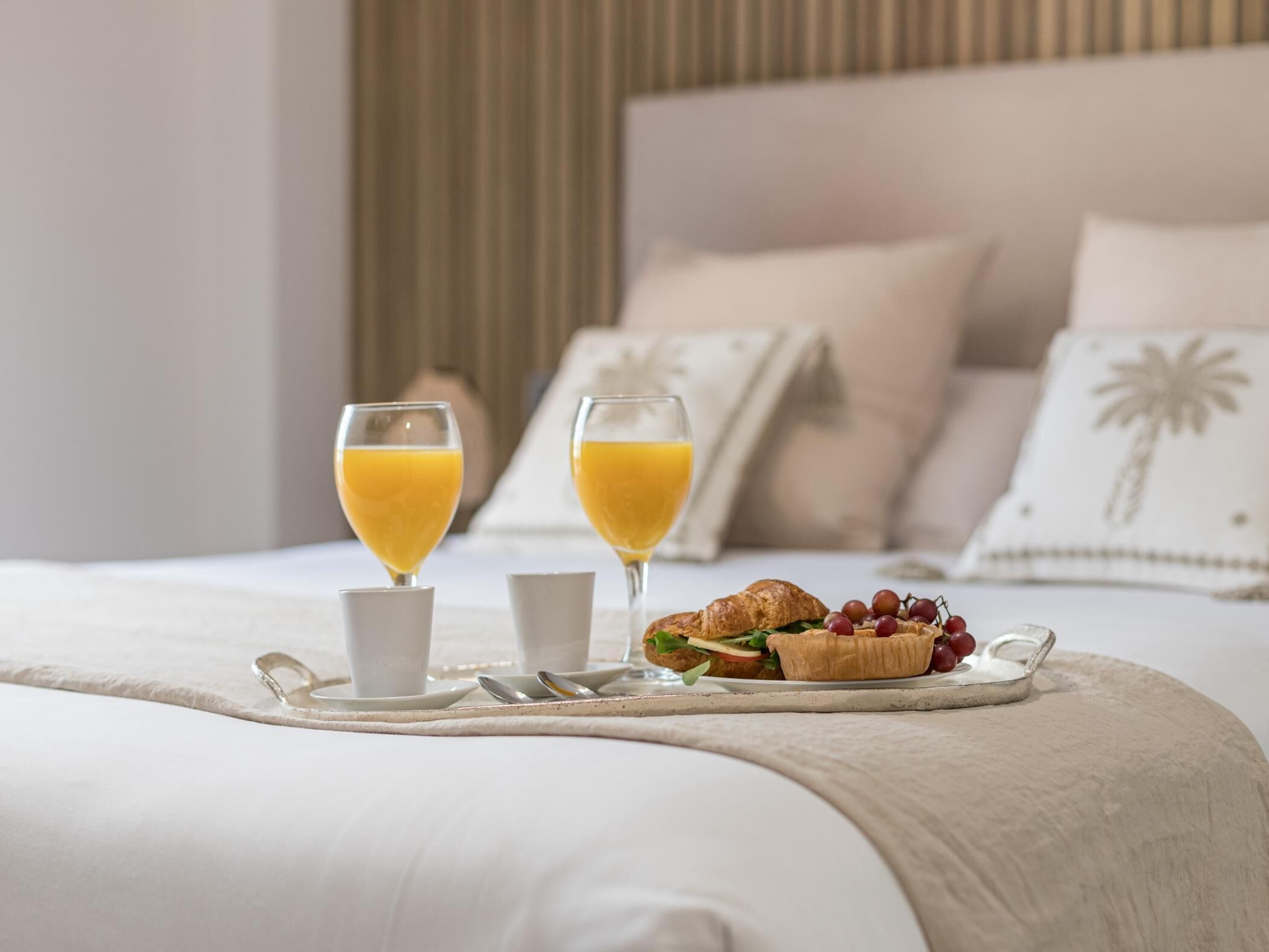 A neatly made breakfast tray on a bed with two glasses of orange juice, a croissant, and a small bowl of grapes.