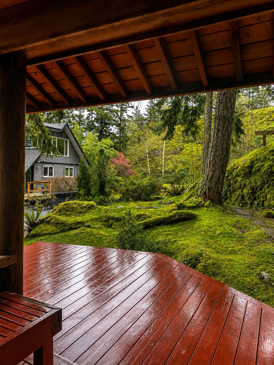 A wooden deck with a bench, overlooking a lush garden with a house in the background.