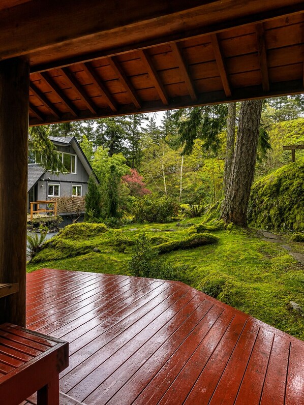 A wooden deck with a bench, overlooking a lush garden with a house in the background.