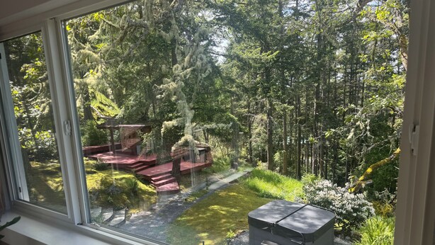 A view through a window of a forested area with a wooden deck and a small shed. - Private Cabin w/ Hot Tub   • Near Beaches & Trails 30 min from Victoria • King Bed (Victoria)