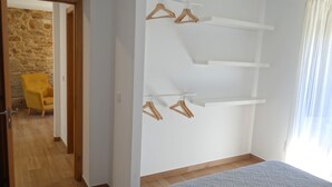A bedroom with a wooden floor, a bed with a grey quilt, and a white wall with three shelves and two wooden hangers.
