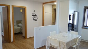 A room with a dining table, chairs, a kitchen area with a refrigerator and oven, and a bedroom visible through an open door.