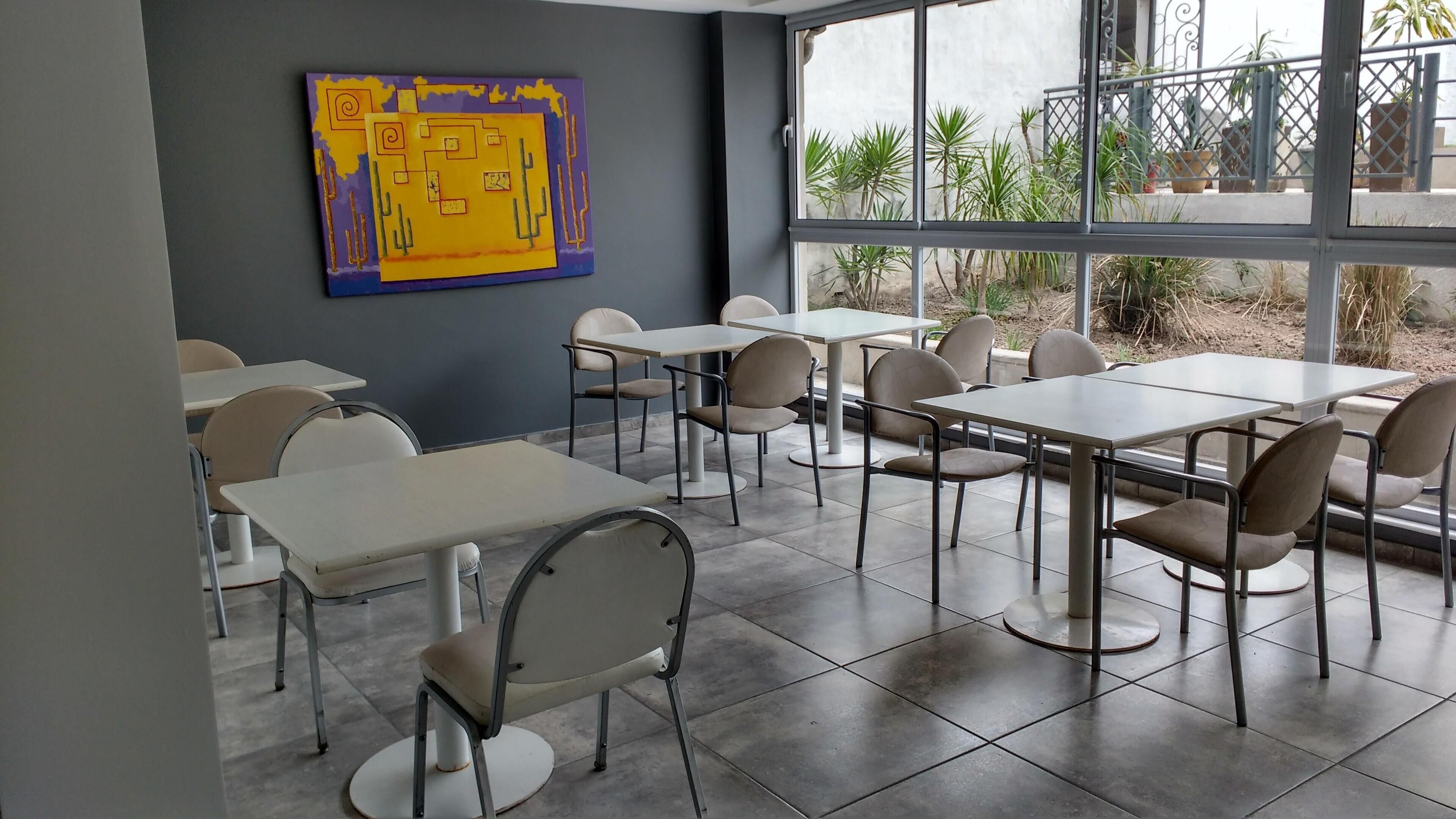 Un espace repas moderne, avec des tables et des chaises, une grande fenêtre donnant sur des plantes et un tableau abstrait aux couleurs vives accroché au mur.