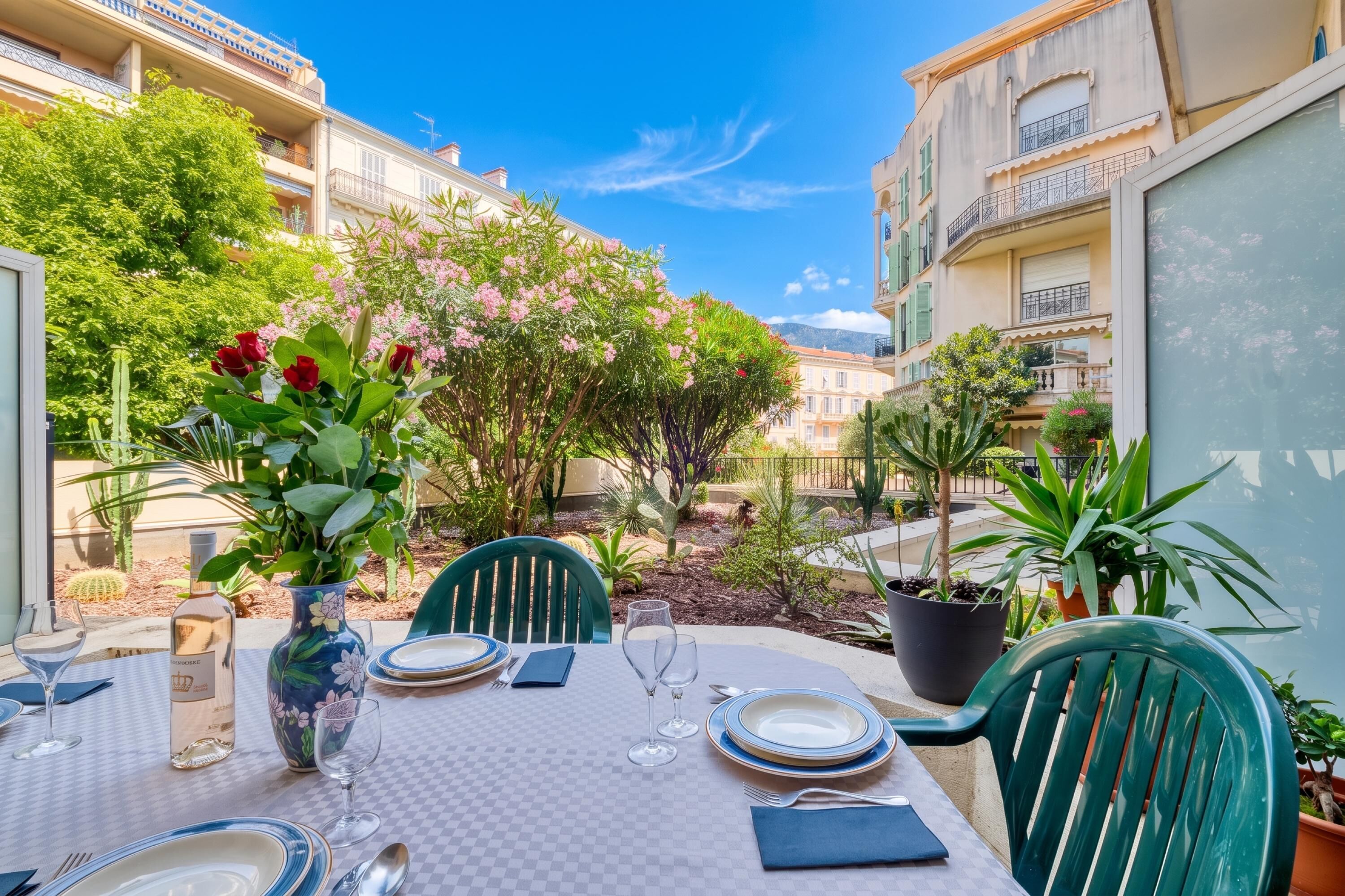 Un espace de restauration en plein air avec une table dressée pour deux personnes, entouré de plantes en pot et offrant une vue sur des immeubles d’habitation.