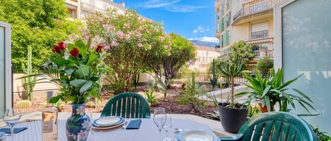 An outdoor dining area with a table set for two, surrounded by potted plants and a view of residential buildings.