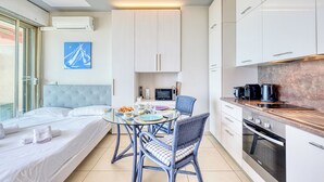 Ein modernes Hotelzimmer mit Bett, einem Glastisch, zwei Stühlen, einer Küchenzeile mit Geräten und Blick ins Freie.