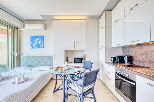 A modern hotel room with a bed, a glass dining table, two chairs, a kitchen area with appliances, and a view of the outside. - Garavan Studio 13 with Sea View Terrace, Menton, France (Menton)