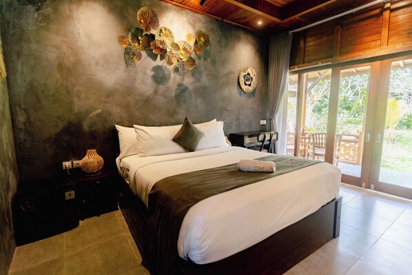 A bedroom with a large bed, a nightstand, a lamp, and a wall decoration. - Puri Kurnia Ayu (Payangan)