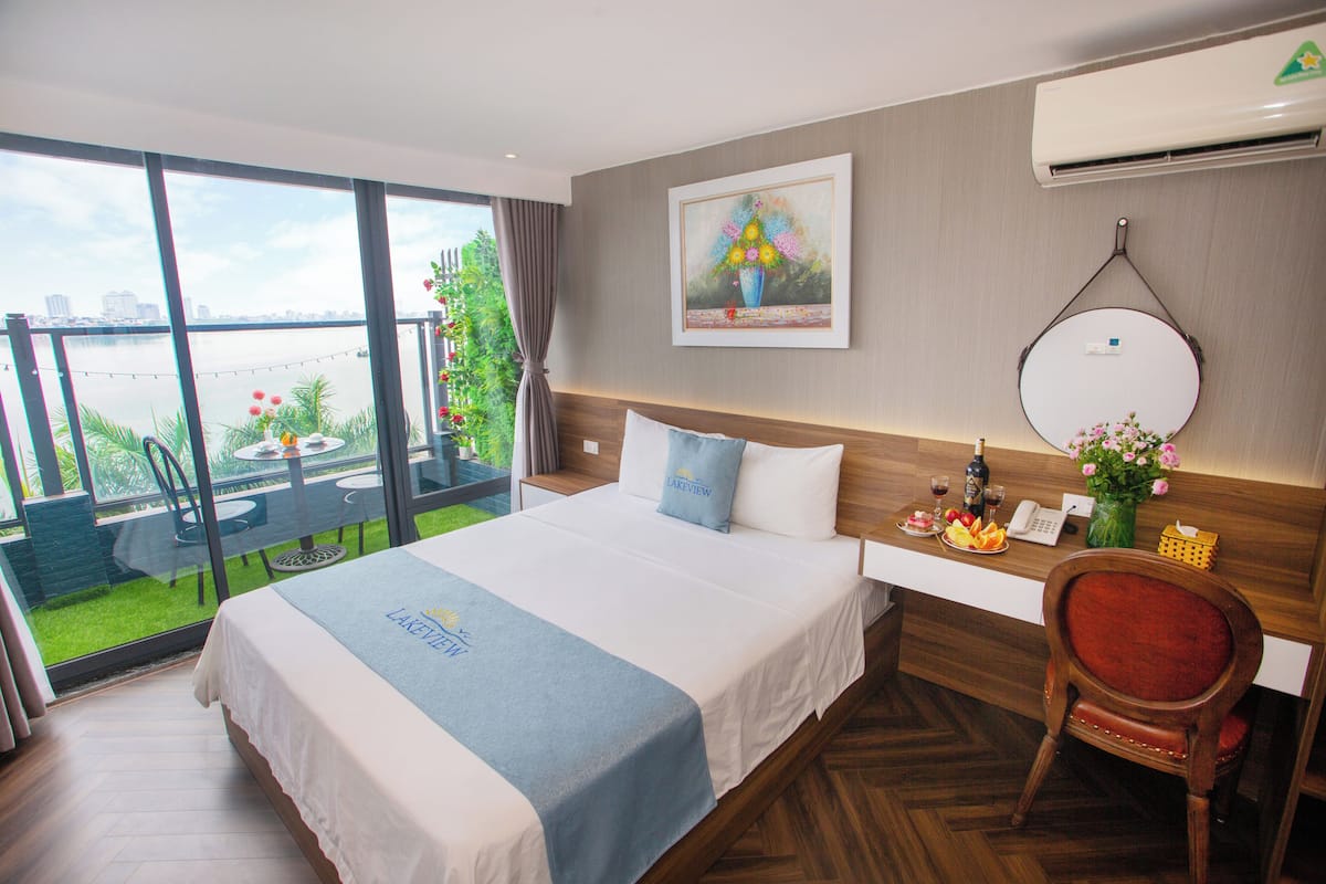A hotel room with a large bed, a desk with a telephone, a chair, and a view of a balcony with a table and chairs.