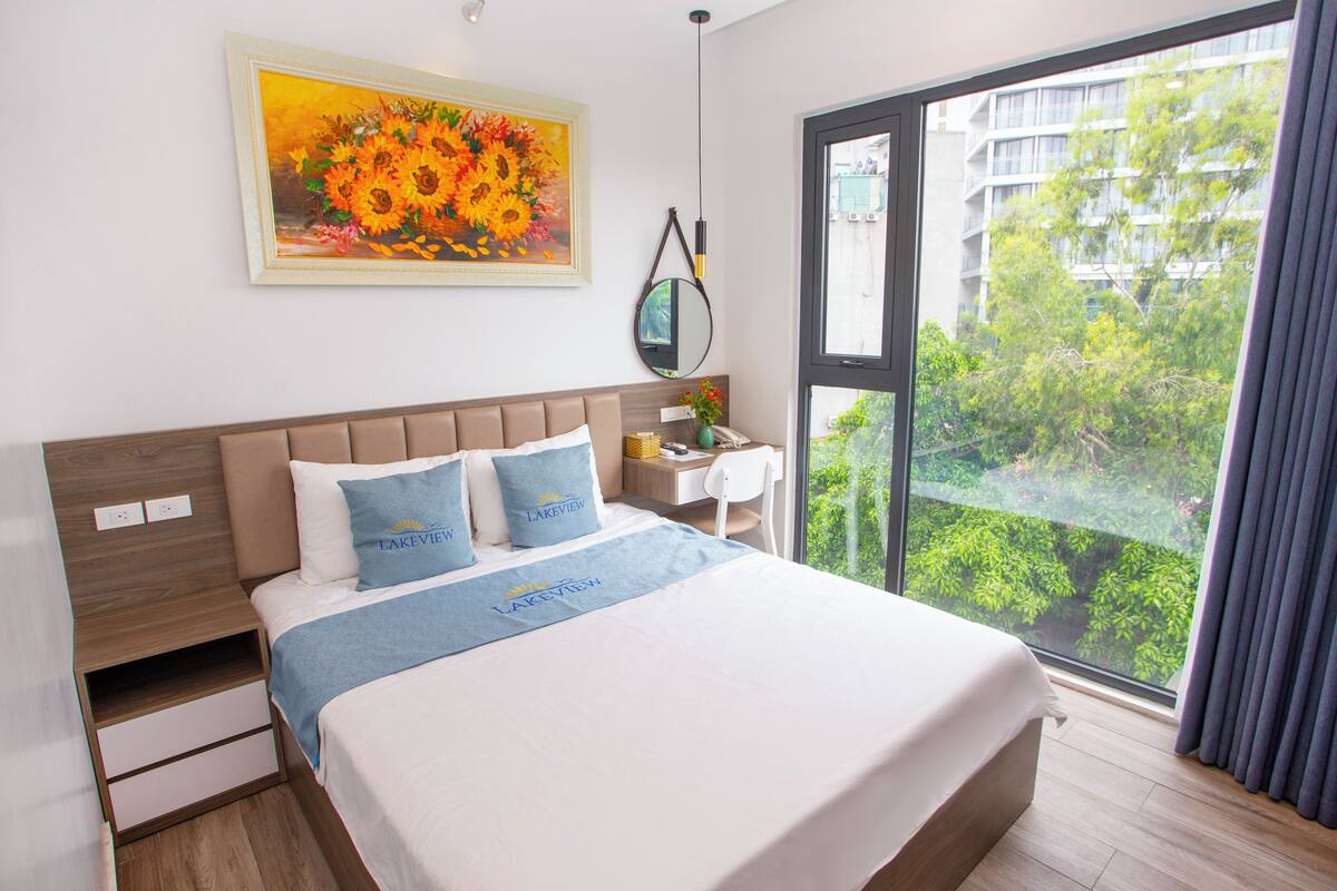 A modern hotel room with a large bed, a nightstand, a desk, and a window with a view of greenery.