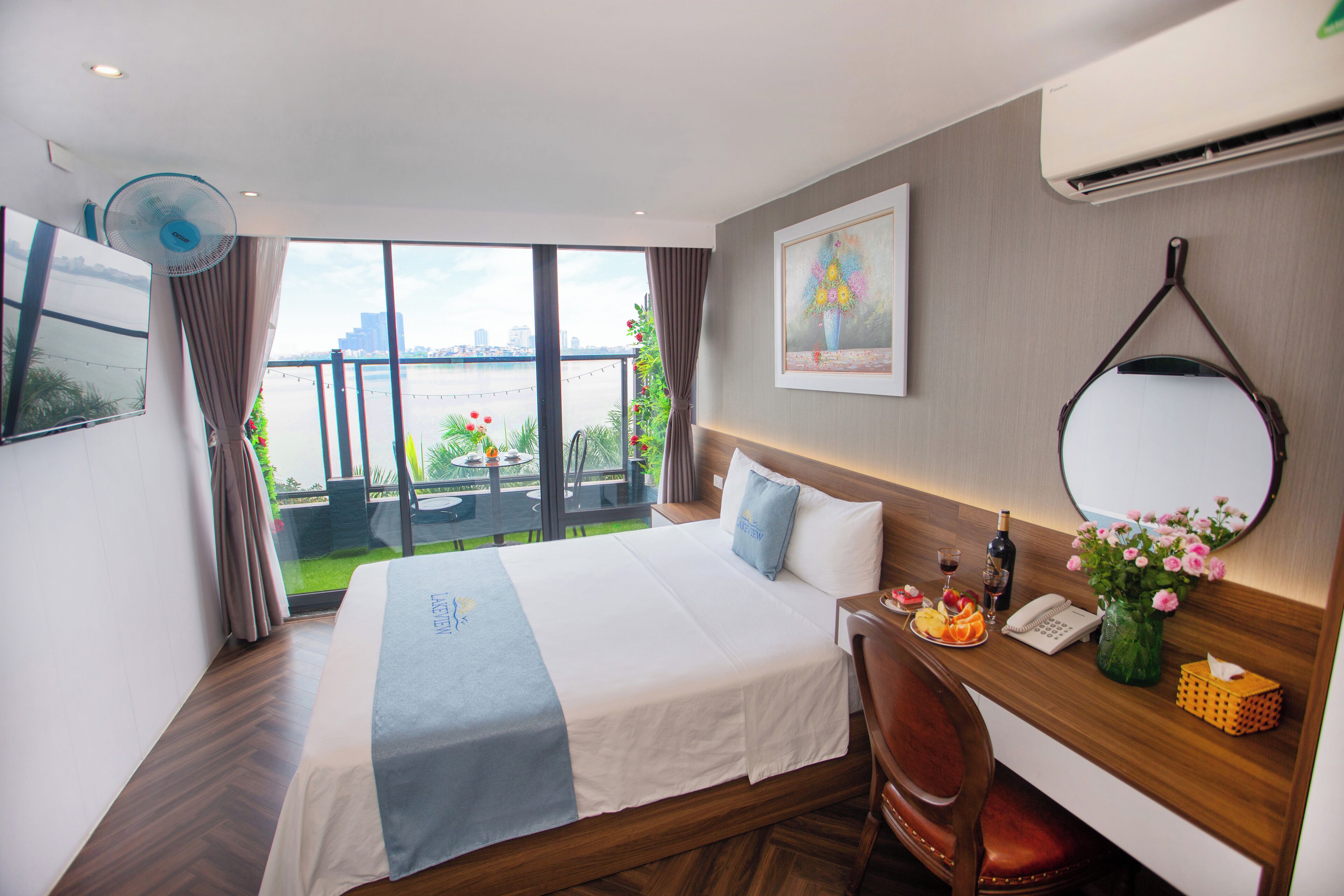 Hotelkamer met een groot bed, een bureau met stoel, een ventilator, een telefoon en uitzicht op de stad en het water.