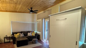 A room with a wooden ceiling, a ceiling fan, a sofa with cushions, a large white wardrobe, and a sliding glass door leading to a balcony. - Rustic, Artsy Studio w/ Desert Panoramic Views  (Black Canyon City)