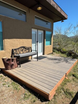 A wooden deck with a bench and a tree stump, a building with a sliding glass door, and a clear view of the surrounding landscape. - Rustic, Artsy Studio w/ Desert Panoramic Views (Black Canyon City)