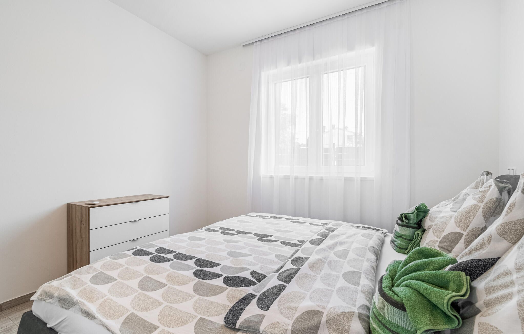 A bedroom with a bed, a bedside table, and a window with sheer curtains.
