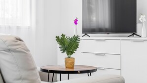 En moderne stue med et fladskærms-tv på et hvidt skab, et rundt træbord med en vævet plante, og en beige sofa.