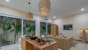 A modern living room with a wooden dining table, a beige sofa, and a large window with tropical plants. - Villa Palima (Canggu)