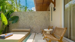 A sunlit outdoor relaxation area with a stone wall, a cushioned bench, wicker chairs, and a small table. - Villa Palima (Canggu)