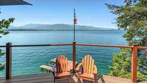 Una terraza de madera con dos sillas Adirondack que dan a un lago tranquilo con montañas al fondo.