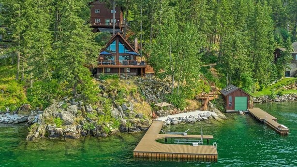 Una cabaña con terraza de madera y un muelle que se adentra en un lago, rodeada de un bosque frondoso.