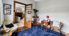Een kamer met een blauw pluisje tapijt, een houten deur, een beige fauteuil, een houten bijzettafeltje met een schaal, een klein tafeltje met een lamp en twee rieten stoelen.