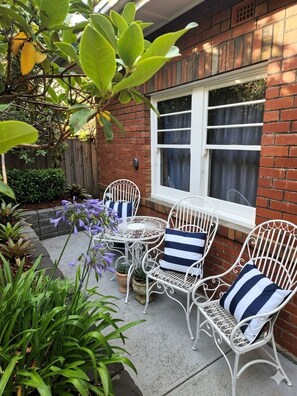 A patio with white metal chairs and a table, surrounded by potted plants and a brick wall.