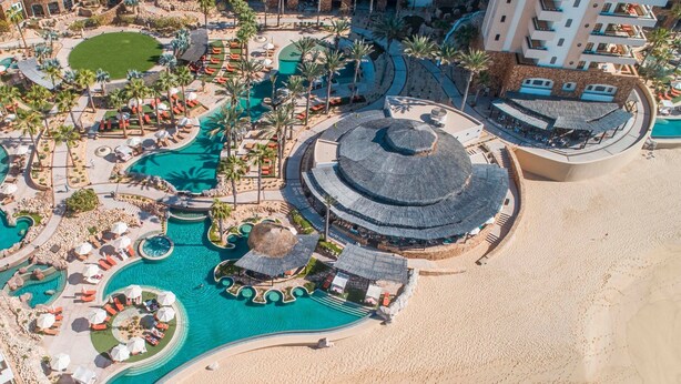 Ovenudblik over et resort med flere swimmingpools, et strandområde og en central bygning med et cirkulært tag.