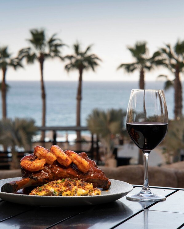 A plate of food with a glass of red wine on a table with a view of the ocean and palm trees. - Cozy resort in amazing Cabo San Lucas with AC, fitness room (Cabo San Lucas)