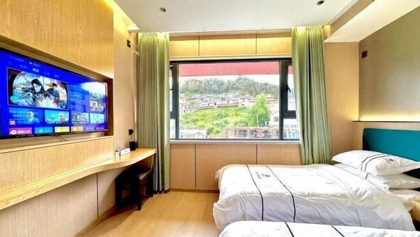 Ein Hotelzimmer mit Fernseher an der Wand, zwei Betten, einem Fenster mit Blick auf Gebäude und einem Vorhang.